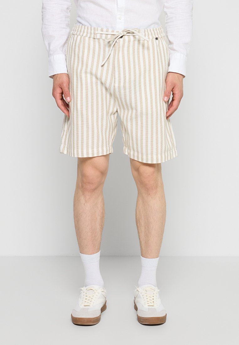 Mann trägt beige und weiß vertikal gestreifte Shorts mit Kordelzug, weiße Socken, weiße Sneaker und ein weißes langärmeliges Hemd und steht.