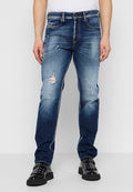 Mann trägt mittelblaue, verwaschene Jeans mit Rissen an den Oberschenkeln und schwarze Lederschuhe mit metallischen Ösen und Schnallen.