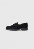 Schwarzer Wildleder-Slipper mit abgerundetem Zehenbereich, flacher Gummisohle und einem dekorativen Riemen über dem Spann. Einfaches, minimalistisches Design.