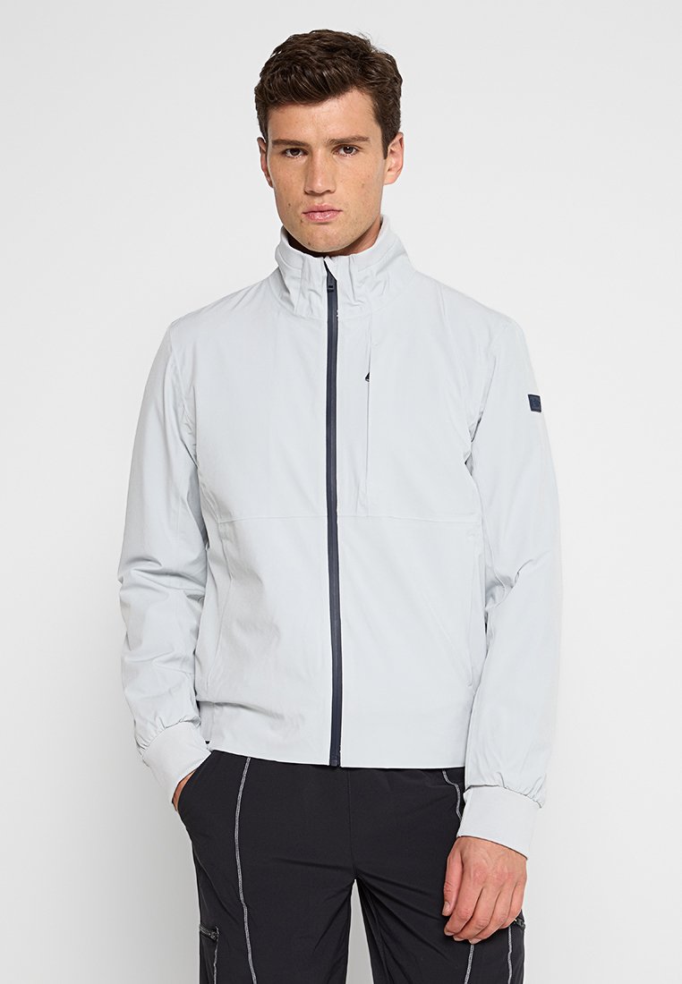 Hellgraue Zip-Jacke mit hohem Kragen, elastischen Bündchen und schwarzen Akzenten; aus leichtem, glattem Material gefertigt.