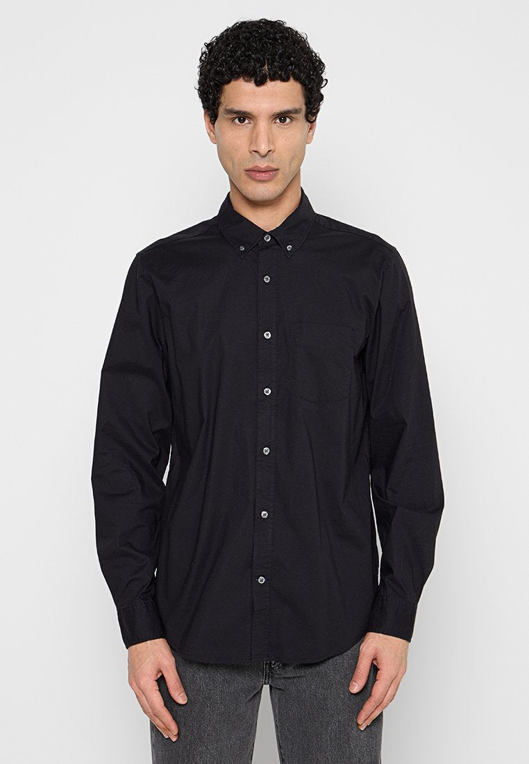 Mann mit lockigem Haar, der ein schwarzes, geknöpftes Langarmhemd und dunkelgraue Jeans trägt, vor einem schlichten hellen Hintergrund stehend.