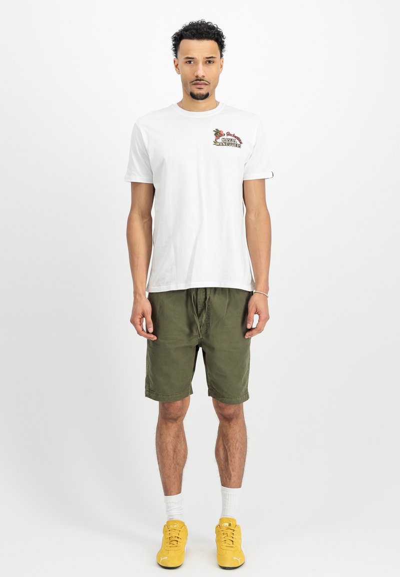 Weißes Baumwoll-T-Shirt mit gesticktem Design, kombiniert mit olivgrünen Shorts. Das Model trägt gelbe Sneakers und weiße Socken.
