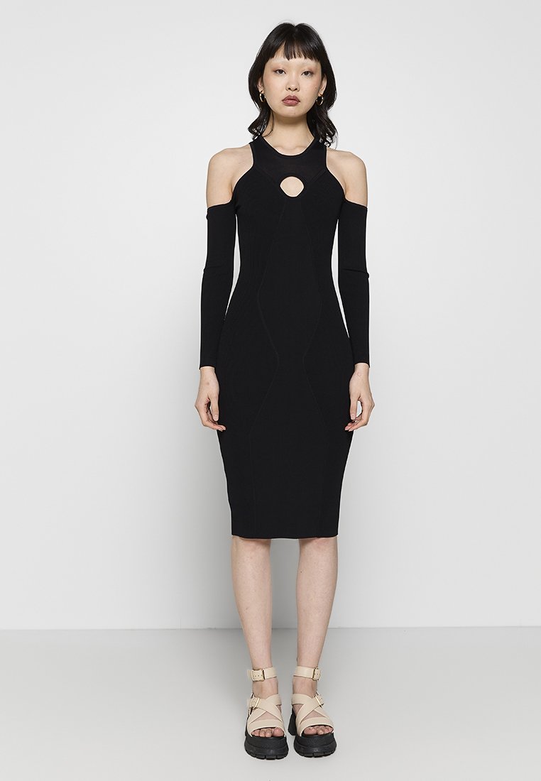 Frau in einem schwarzen Midi-Kleid mit kalten Schultern und einem herzförmigen Ausschnitt sowie beigen Plateausandalen, die vor einem weißen Hintergrund steht.