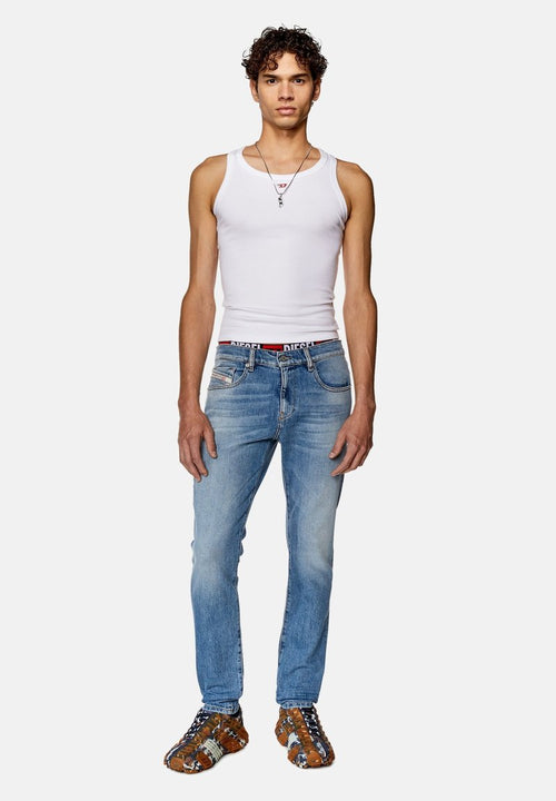 Schmal geschnittene blaue Jeans mit leichtem Waschung, ausgestattet mit einem gebrandeten Bund. Kombiniert mit einem weißen Tanktop und gemusterten Schuhen.