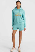 Türkise Kapuzenjacke mit goldener Grafik, kombiniert mit passenden Shorts. Weiches Material, lockere Passform und Kängurutasche. Beigefarbene und rosafarbene Sneakers.