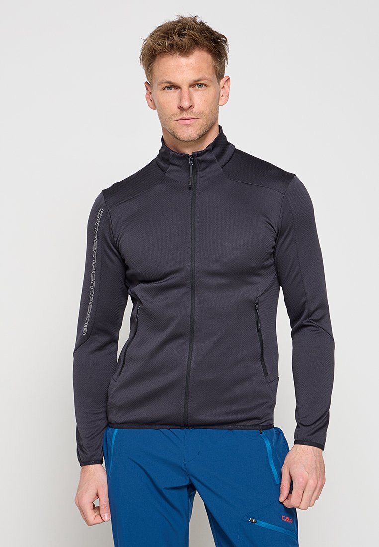 Mann trägt schwarze Zip-up-Jacke und blaue Outdoor-Hose, steht vor einfachem hellem Hintergrund.