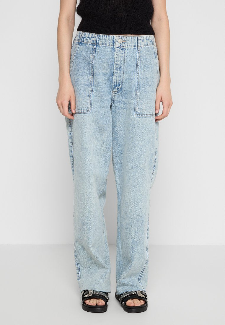 Person, der hellblaue, weite Jeans mit vorderen Taschen trägt, kombiniert mit einem schwarzen ärmellosen Oberteil und schwarzen Sandalen mit Schnallen.