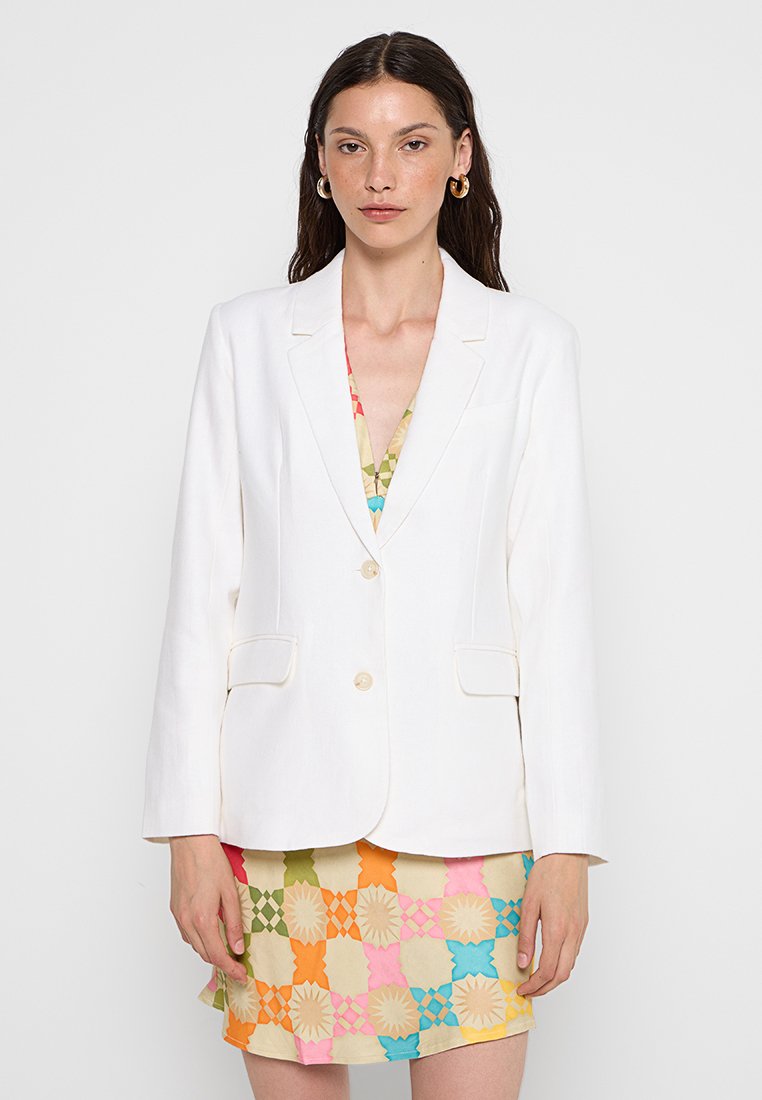Weißer Leinenblazer mit taillierter Passform, mit Reverskragen, zwei vorderen Taschen und einem einzelnen Knopfverschluss, kombiniert mit einem gemusterten Kleid darunter.