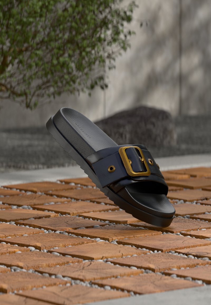 Schwarze Slidesandale mit strukturierter Sohle und breitem Riemen, der eine große goldene Schnalle trägt, liegt auf Holzkacheln im Freien.