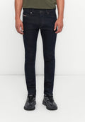 Mann in dunklen Skinny-Jeans, schwarzen und grauen Sneakers und einem grün eingesteckten Oberteil, steht vor einem weißen Hintergrund.