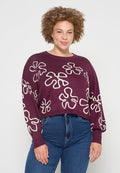 Bordeauxfarbener verkürzter Pullover mit großen weißen Blumenmustern, runder Halsauschnitt und langen Ärmeln, aus weichem Strickmaterial. Sichtbare Jeans aus Denim.