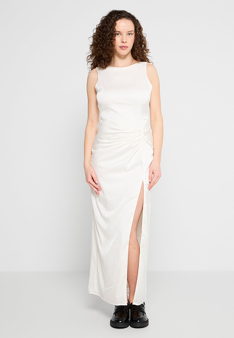 Weißes Satin-Maxikleid mit ärmellosem Design, Knoten-Detail an der Taille und einem seitlichen Schlitz. Das Model trägt schwarze Schuhe.