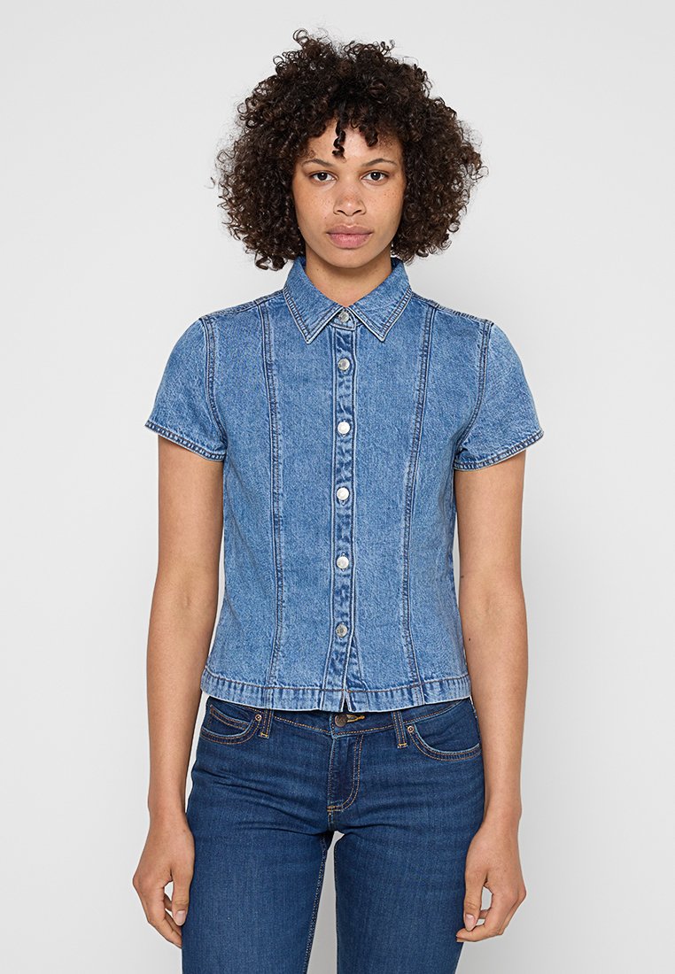 Frau mit lockigem Haar, die ein kurzärmliges, blaues Denim-Hemd mit Knöpfen und dunkelblaue Jeans trägt, steht vor einem schlichten hellen Hintergrund.
