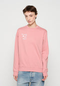 Junge Person trägt ein pinkes Sweatshirt mit dem Schriftzug „Women of the Wave“ und hellblaue Jeans, steht vor einem schlichten hellen Hintergrund.