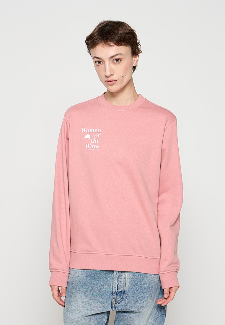 Junge Person trägt ein pinkes Sweatshirt mit dem Schriftzug „Women of the Wave“ und hellblaue Jeans, steht vor einem schlichten hellen Hintergrund.