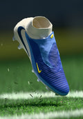 Blauer Fußballschuh mit strukturiertem Obermaterial, weißem Kragen, gelben Akzenten und einem stromlinienförmigen Design, schwebend über Gras mit Schutt.