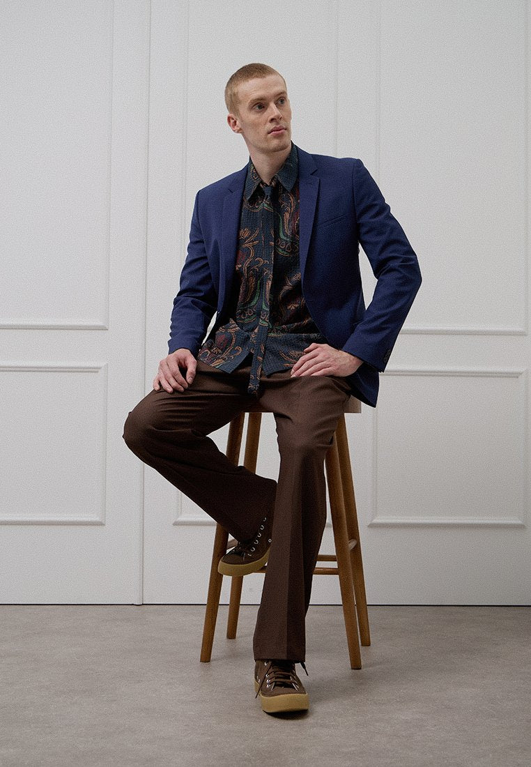 Mann sitzt auf einem hölzernen Hocker, trägt ein blaues Sakko, ein gemustertes Hemd und braune Hosen. Das Hemd zeigt komplexe Designs in dunklen Farben.