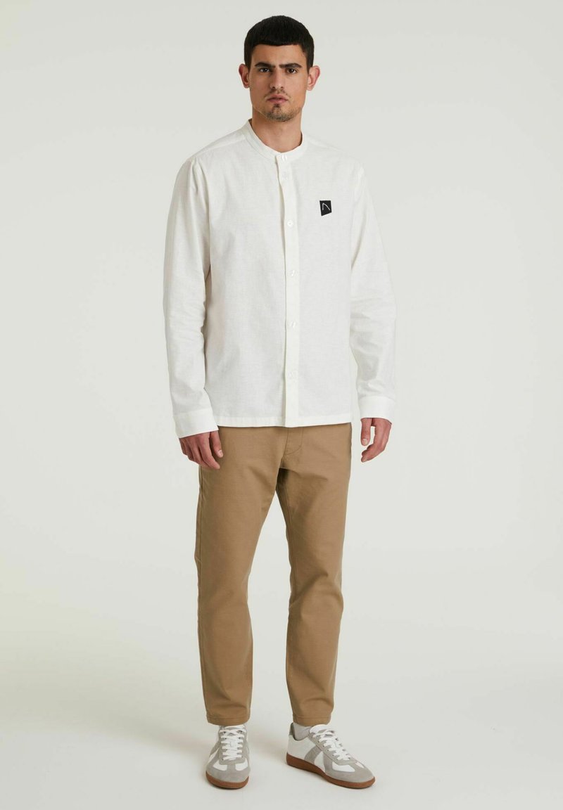 Weißes Hemd mit Stehkragen, schwarzem Logo-Patch, kombiniert mit khakifarbenen Hosen und grauen Sneakers.