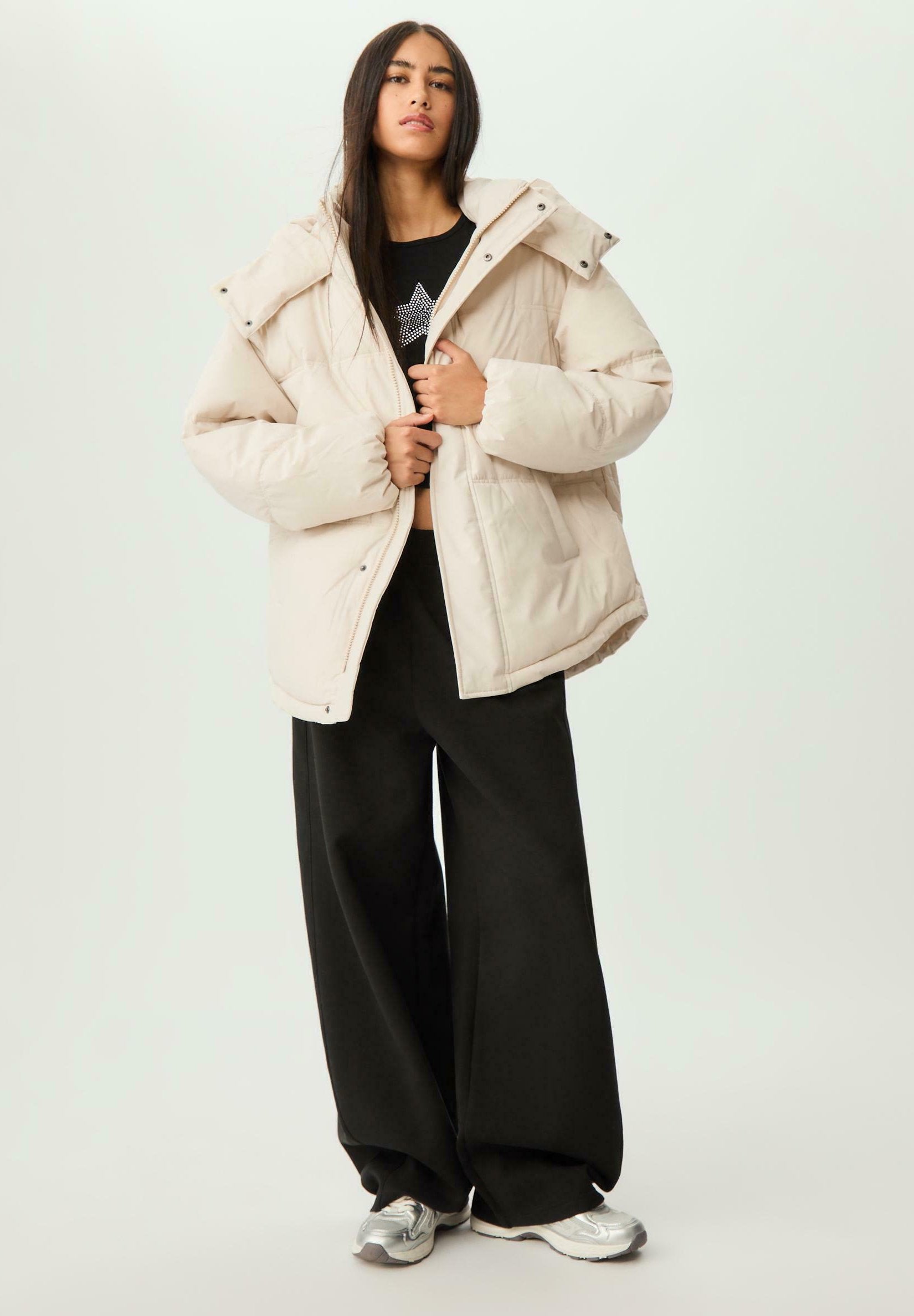 Beige Steppjacke mit hohem Kragen und Reißverschluss, kombiniert mit schwarzen weiten Hosen und silbernen Sneakers. Glatte, gestepfte Textur.