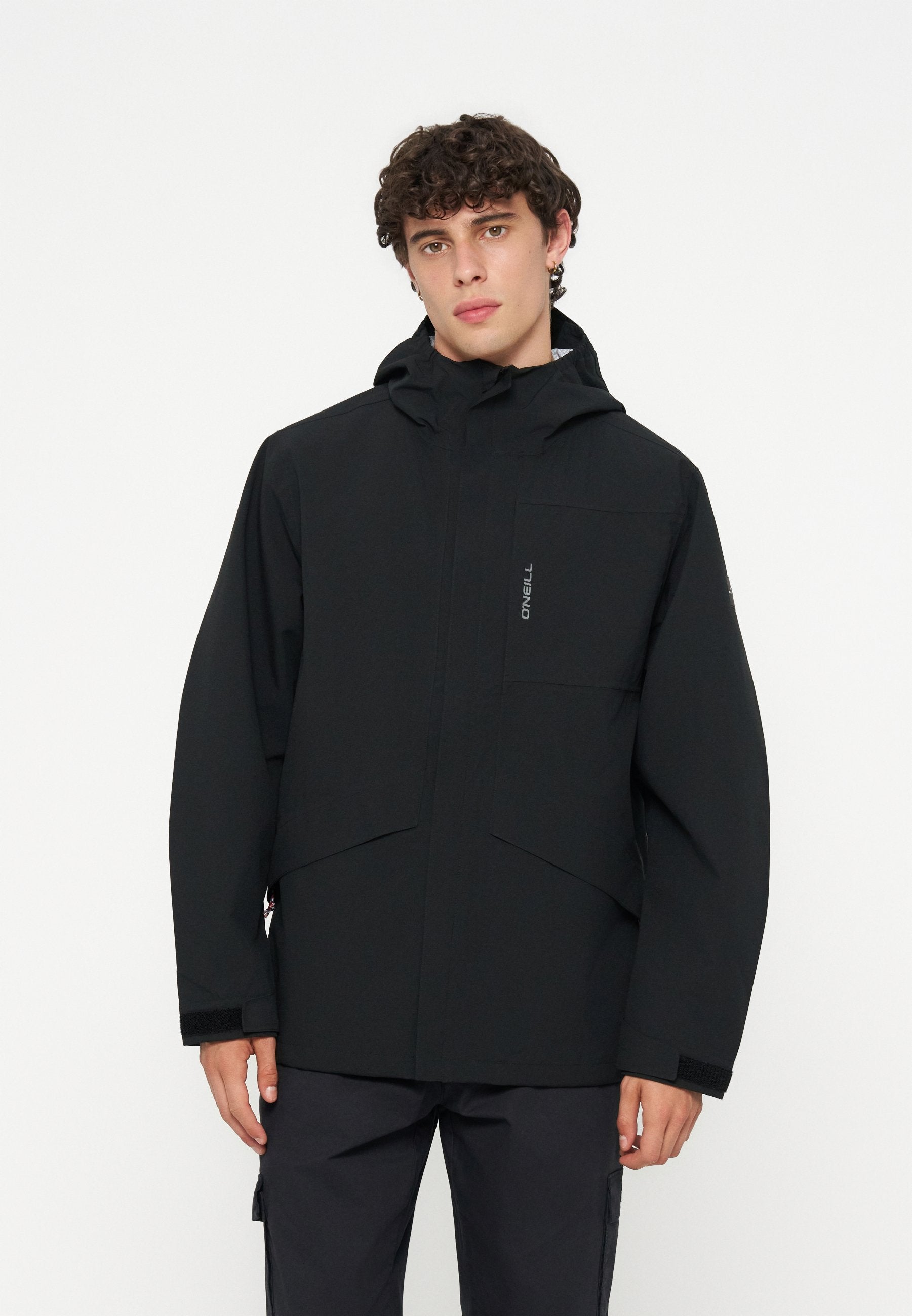 Junger Mann mit lockigem Haar, der eine schwarze Kapuzenjacke mit dem O'Neill-Logo auf der Brusttasche trägt, vor einem einfarbigen Hintergrund stehend.