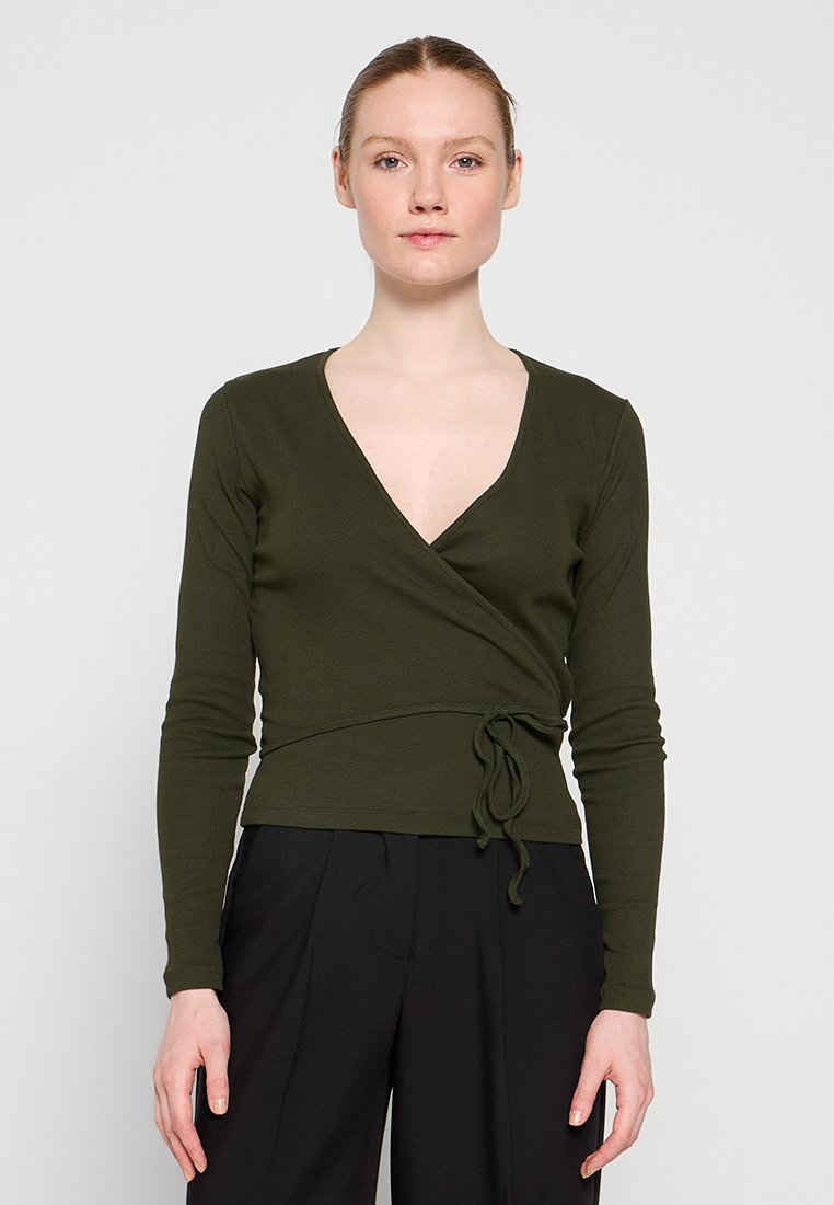 Olivgrünes Wickeloberteil mit langen Ärmeln, V-Ausschnitt, Taillendetail und einem weichen, strukturierten Stoff. Kombiniert mit hochtaillierten schwarzen Hosen.