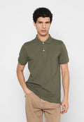 Mann mit lockigem Haar, der ein olivgrünes Poloshirt und beige Hosen trägt, vor einem schlichten hellgrauen Hintergrund stehend.