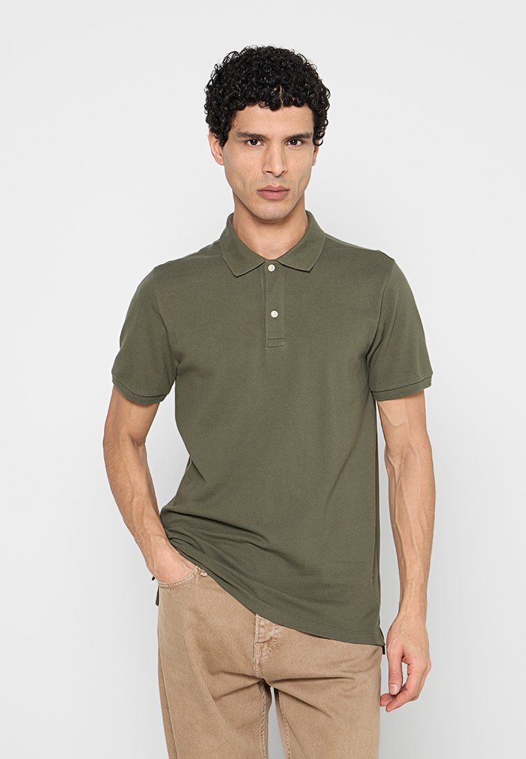 Mann mit lockigem Haar, der ein olivgrünes Poloshirt und beige Hosen trägt, vor einem schlichten hellgrauen Hintergrund stehend.