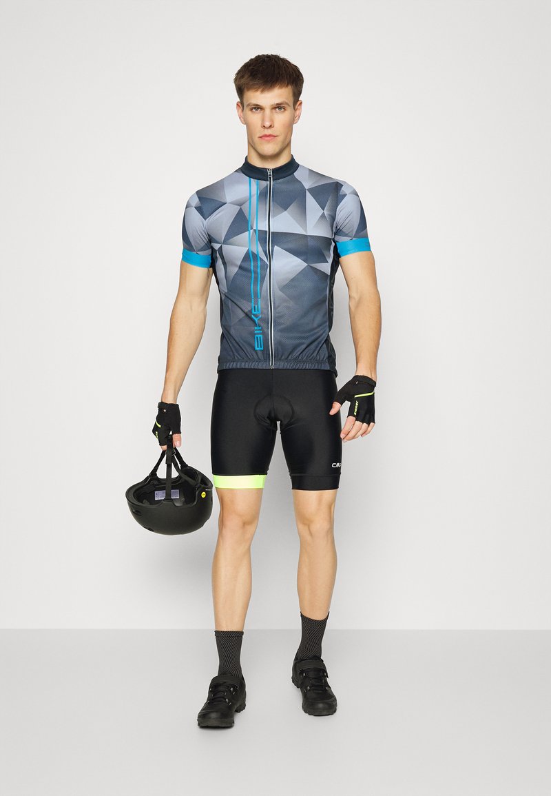 Radbekleidung mit einem grauen geometrischen Muster auf dem Trikot, blauen Akzenten und schwarzen Shorts mit gelbem Saum, ergänzt durch einen schwarzen Helm.