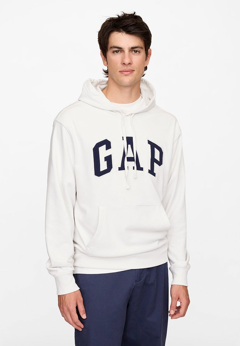 Junger Mann trägt einen weißen GAP-Kapuzenpullover mit marineblauen Hosen, steht vor einem schlichten weißen Hintergrund und blickt nach vorne.