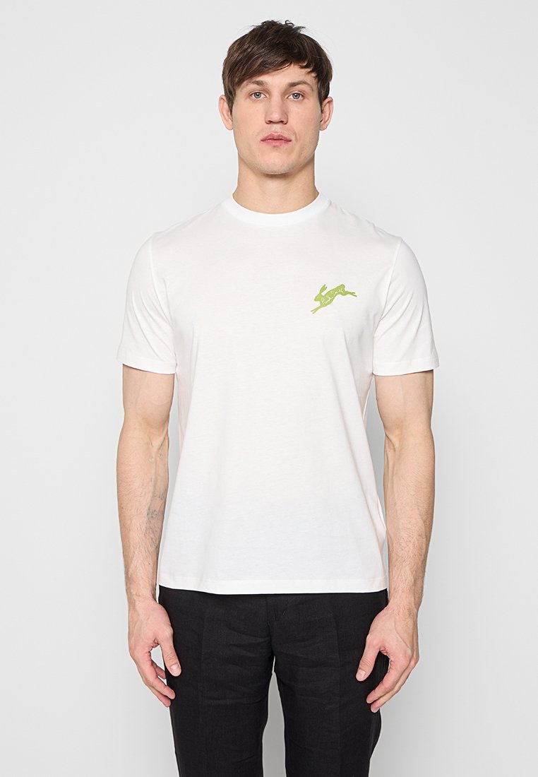 Mann steht frontal, trägt ein schlichtes weißes T-Shirt mit kurzen Ärmeln und einem kleinen grünen Hasen-Logo auf der linken Brust sowie schwarze Hosen.