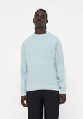 Mann steht und trägt ein hellblaues Sweatshirt mit langen Ärmeln und subtilen Nähten an den Ärmeln sowie schwarze Hosen vor einem schlichten weißen Hintergrund.