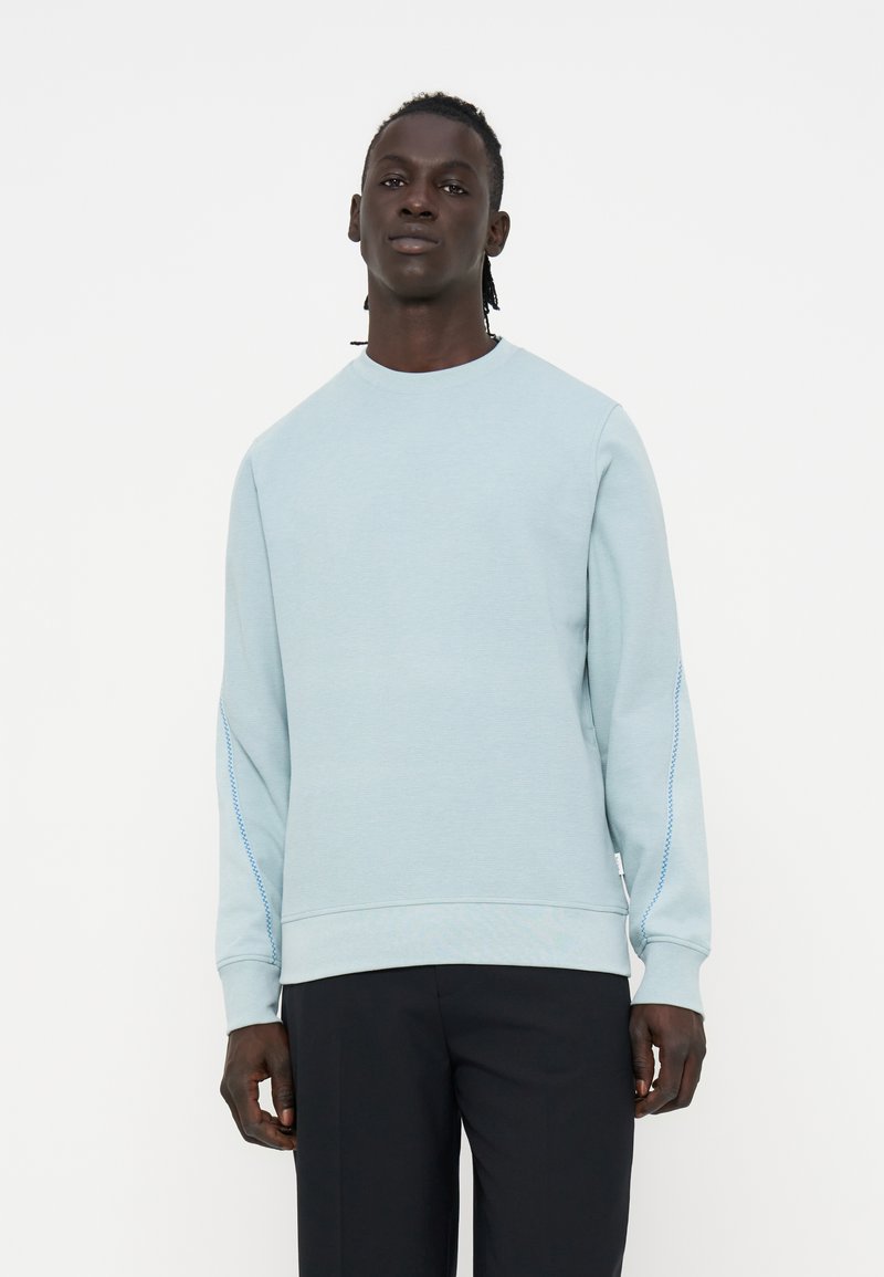 Mann steht und trägt ein hellblaues Sweatshirt mit langen Ärmeln und subtilen Nähten an den Ärmeln sowie schwarze Hosen vor einem schlichten weißen Hintergrund.