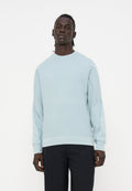 Mann steht und trägt ein hellblaues Sweatshirt mit langen Ärmeln und subtilen Nähten an den Ärmeln sowie schwarze Hosen vor einem schlichten weißen Hintergrund.