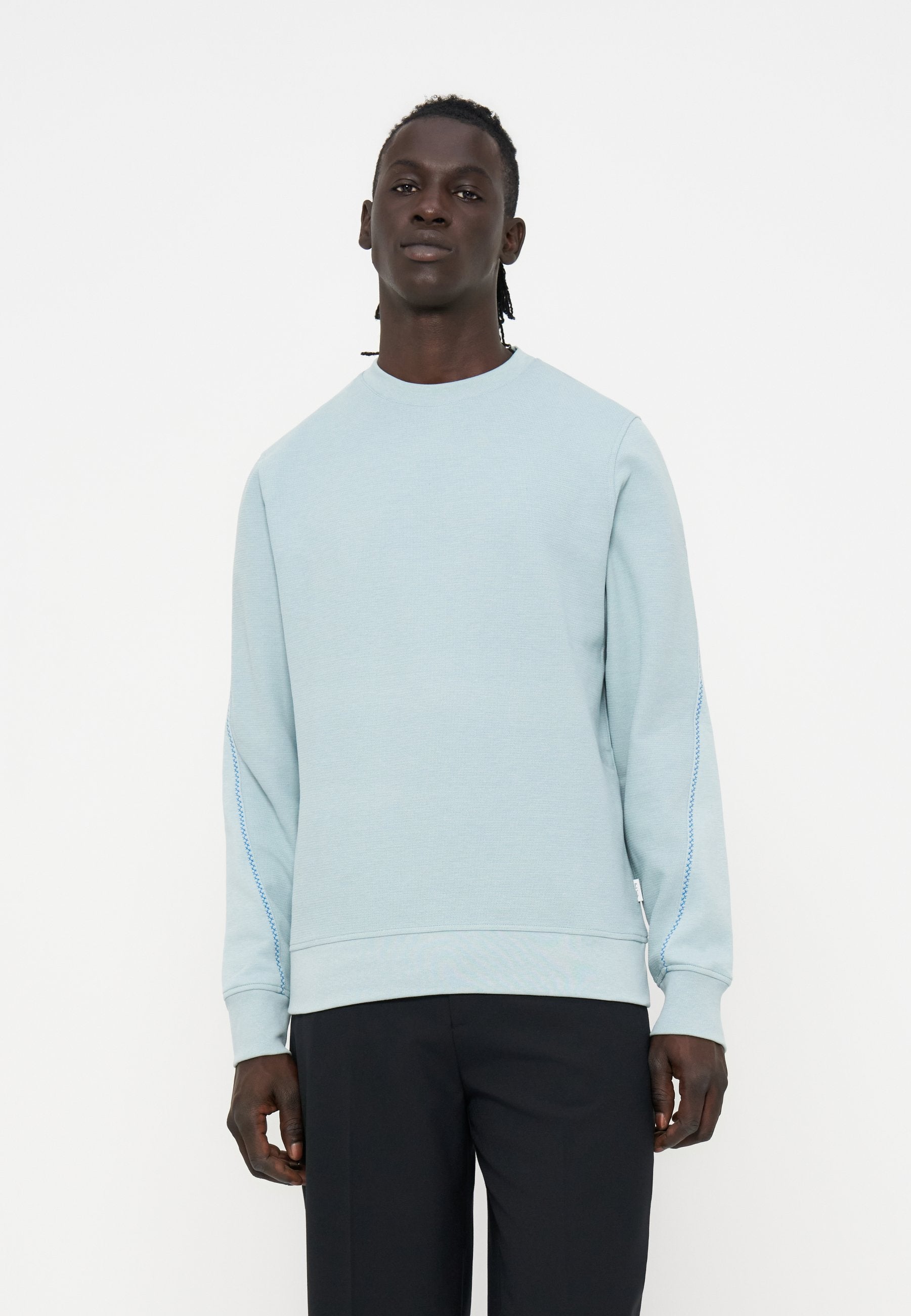 Mann steht und trägt ein hellblaues Sweatshirt mit langen Ärmeln und subtilen Nähten an den Ärmeln sowie schwarze Hosen vor einem schlichten weißen Hintergrund.