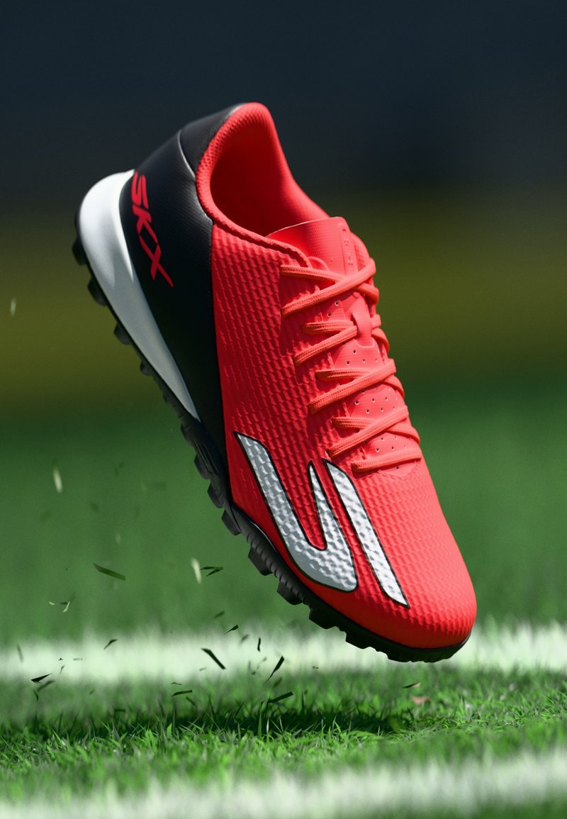 Leuchtend roter Fußballschuh mit schwarzer Ferse und weißer Außensohle, mit der Marke "SKX", schwebt über Gras, aus dem kleine Halme verstreut werden.