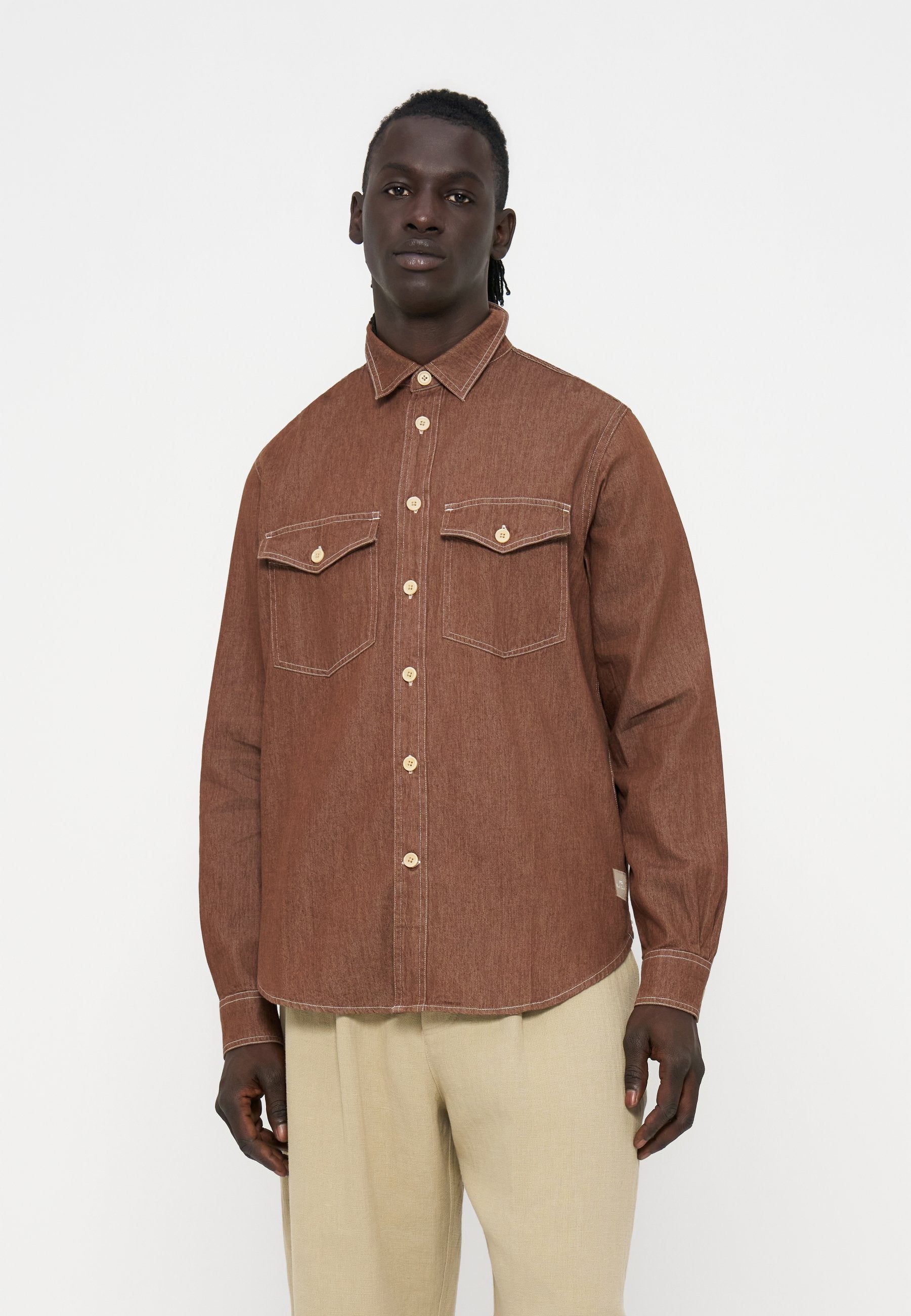 Mann trägt ein braunes Hemd mit Knöpfen und zwei Brusttaschen sowie beige Hose und steht vor einem schlichten weißen Hintergrund.