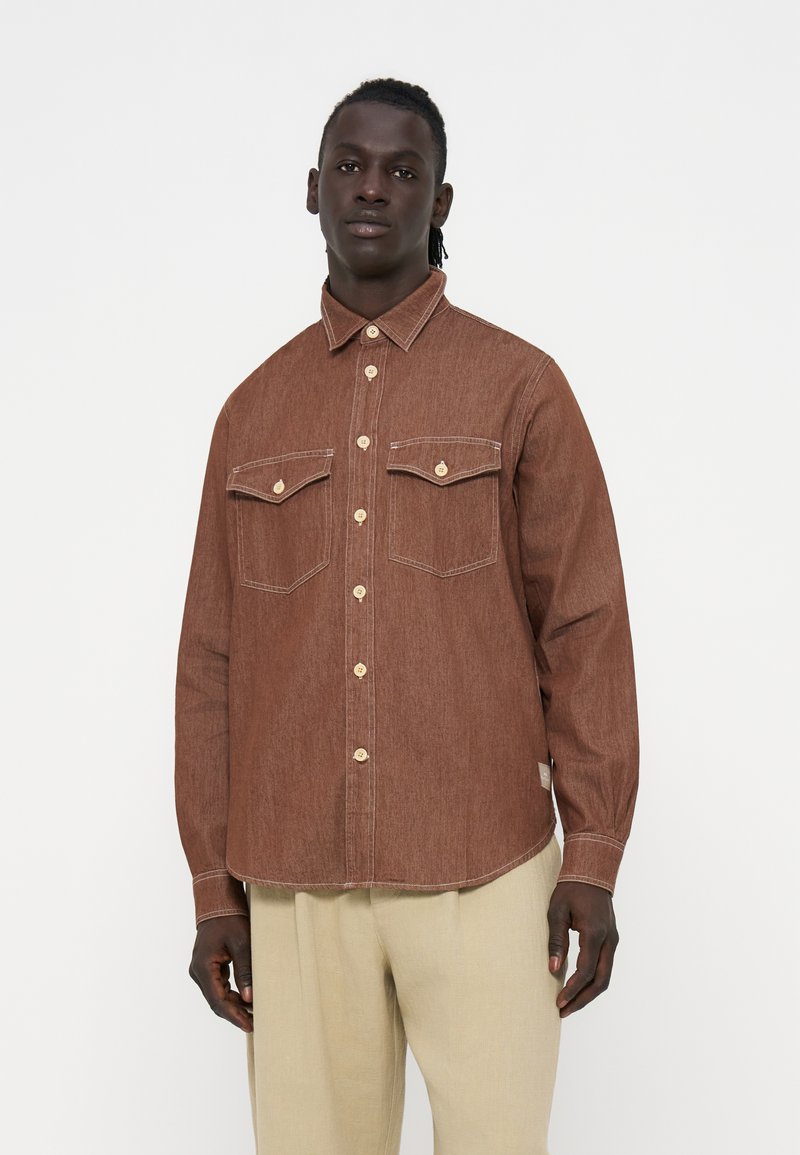 Mann trägt ein braunes Hemd mit Knöpfen und zwei Brusttaschen sowie beige Hose und steht vor einem schlichten weißen Hintergrund.