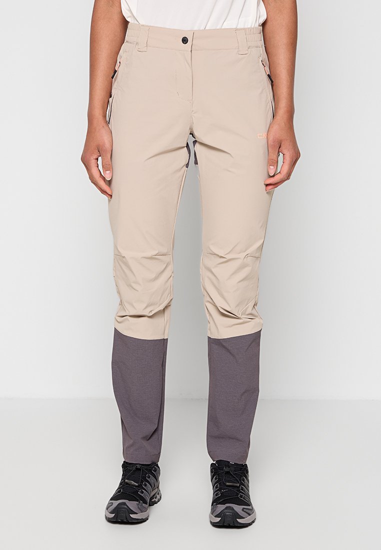 Person trägt beige und graue Outdoor-Hose und schwarze Wanderschuhe und steht vor einem schlichten weißen Hintergrund.