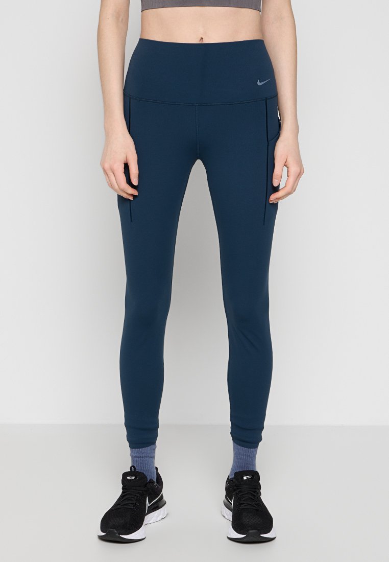Person, der navyblaue, hoch taillierte Leggings mit seitlichen Taschen, graue Socken bis zum Knöchel und schwarze Nike-Laufschuhe trägt, steht vor einem weißen Hintergrund.