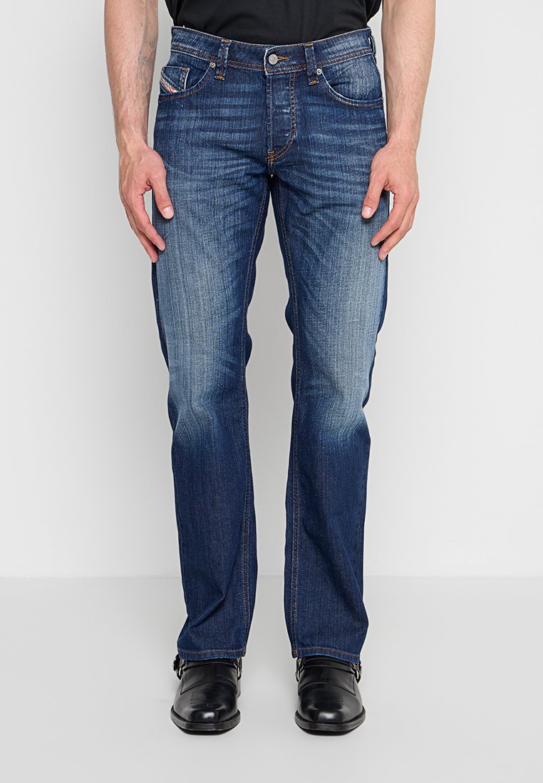 Mann trägt dunkelblaue Jeans mit geradem Bein und verblassten Details, schwarze Lederschuhe und ein schwarzes Hemd vor einem schlichten weißen Hintergrund.