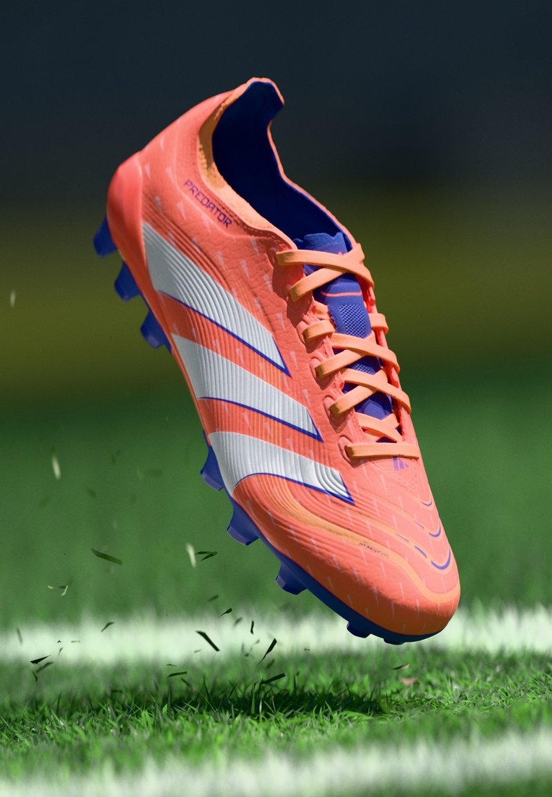 Orange und blaue Fußballschuhe mit strukturiertem Obermaterial, weißen und blauen Streifen sowie lila Stollen, die über grünem Gras schweben.