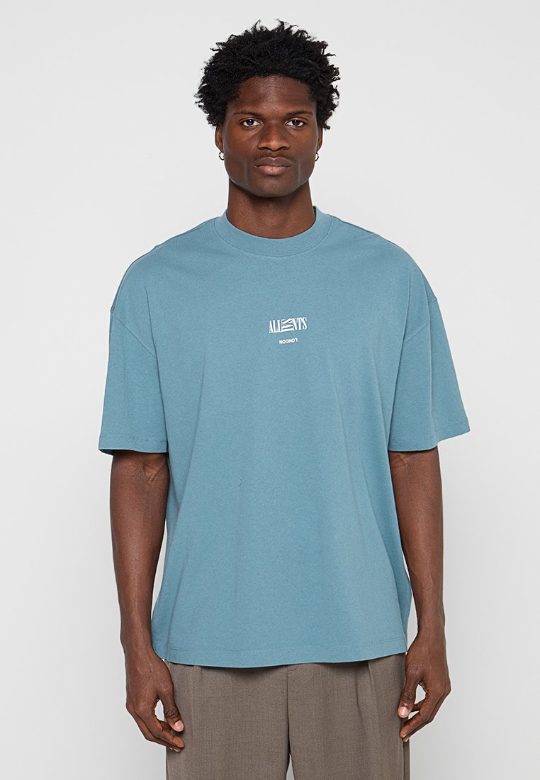 Mann mit blauem übergroßem T-Shirt mit "ALLSAINTS LONDON"-Logo, steht vor einem schlichten hellgrauen Hintergrund.