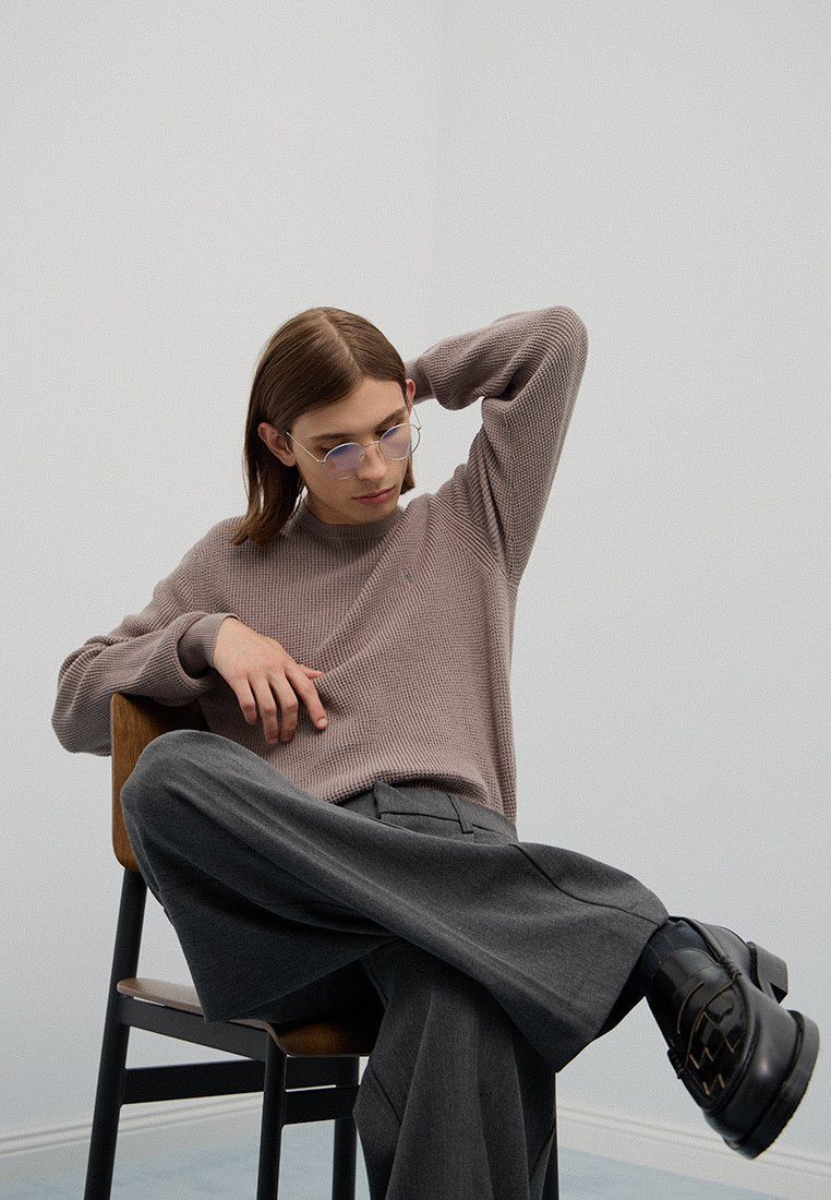 Brauner gerippter Strickpullover, graue weit geschnittene Hose und schwarze Schnürschuhe. Klare Brille mit dünnen Rahmen. Sitzt auf einem Holzstuhl.