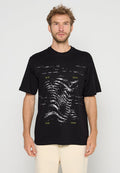 Mann mit kurzen lockigen Haaren, der ein schwarzes Grafik-T-Shirt mit verzerrtem weißen und gelben Textdesign und beigefarbenen Hosen trägt, steht vor einem einfarbigen Hintergrund.
