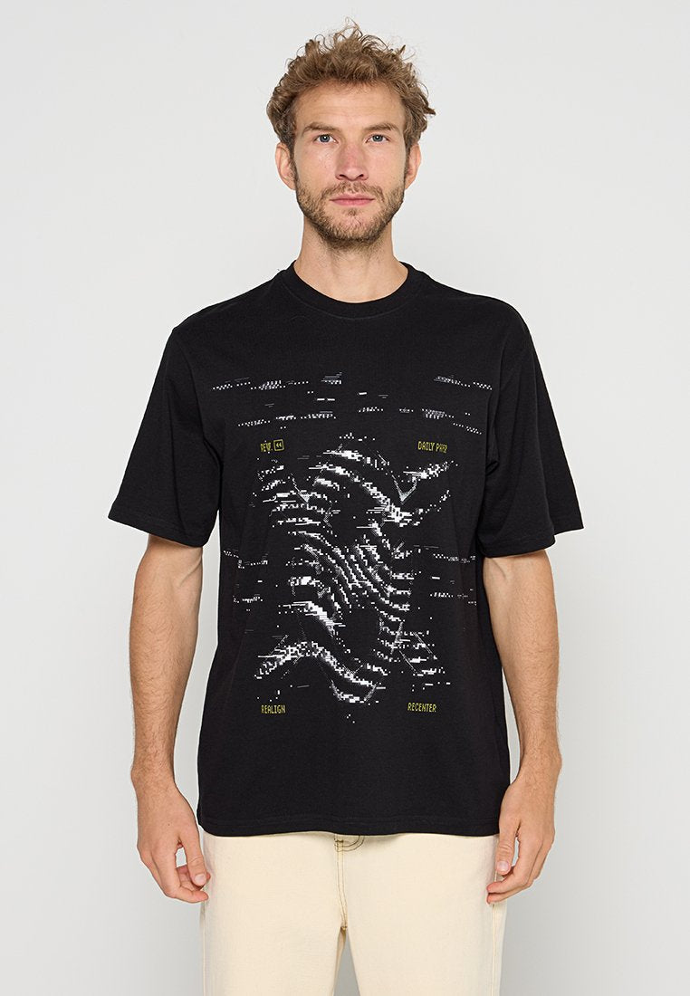 Mann mit kurzen lockigen Haaren, der ein schwarzes Grafik-T-Shirt mit verzerrtem weißen und gelben Textdesign und beigefarbenen Hosen trägt, steht vor einem einfarbigen Hintergrund.