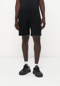 Ein Mann trägt schwarze Shorts mit Kordelzug, weiße Socken und schwarze Sneakers und steht vor einem einfarbigen weißen Hintergrund.