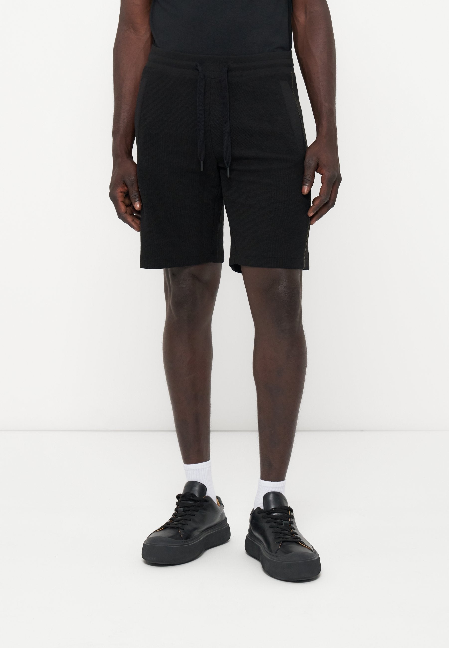 Ein Mann trägt schwarze Shorts mit Kordelzug, weiße Socken und schwarze Sneakers und steht vor einem einfarbigen weißen Hintergrund.
