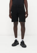 Ein Mann trägt schwarze Shorts mit Kordelzug, weiße Socken und schwarze Sneakers und steht vor einem einfarbigen weißen Hintergrund.