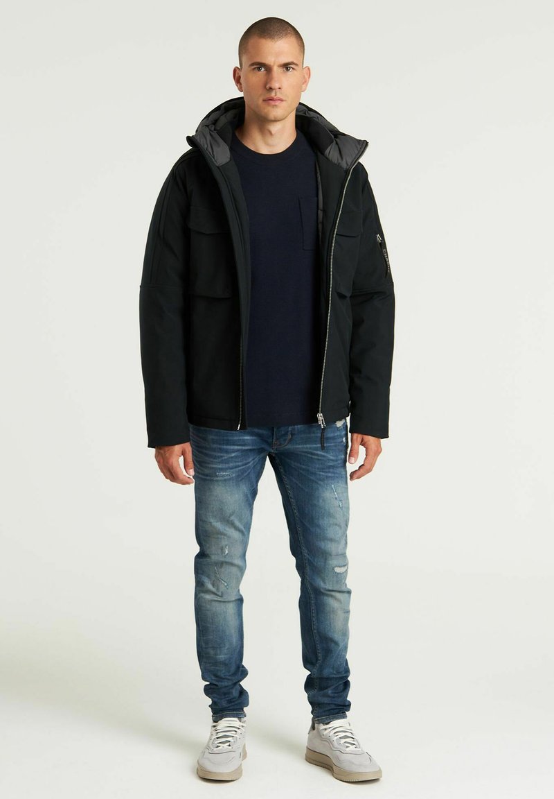Schwarze Kapuzenjacke mit Reißverschluss, mehreren Taschen und grauem Innenfutter. Kombination mit einem marineblauen Shirt, verwaschenen blauen Jeans und Turnschuhen.
