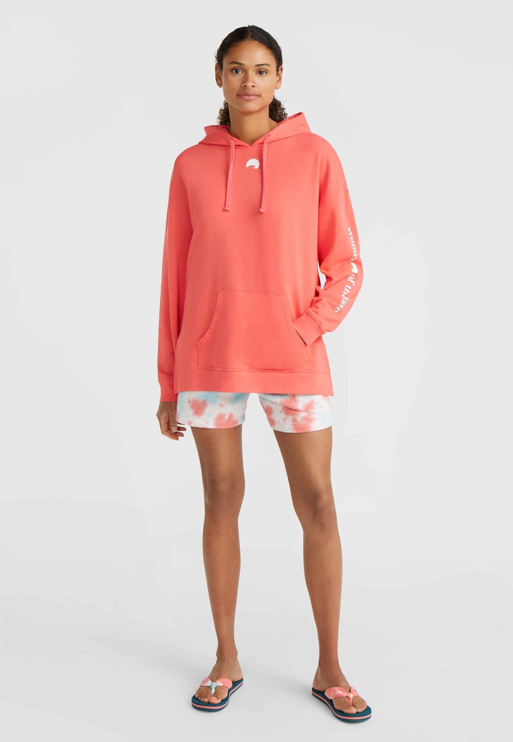 Korallefarbener Hoodie aus weichem Material mit einer Vorderasche, kombiniert mit hellen Batik-Shorts und Flip-Flops mit einem pinken Schleifendetail.